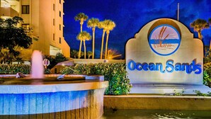 A nighttime view of Ocean Sands hotel with a fountain, palm trees, and a sign with the hotel name.