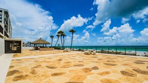 A spacious outdoor area with a stone patio, a thatched-roof structure, palm trees, and a view of the beach and ocean.