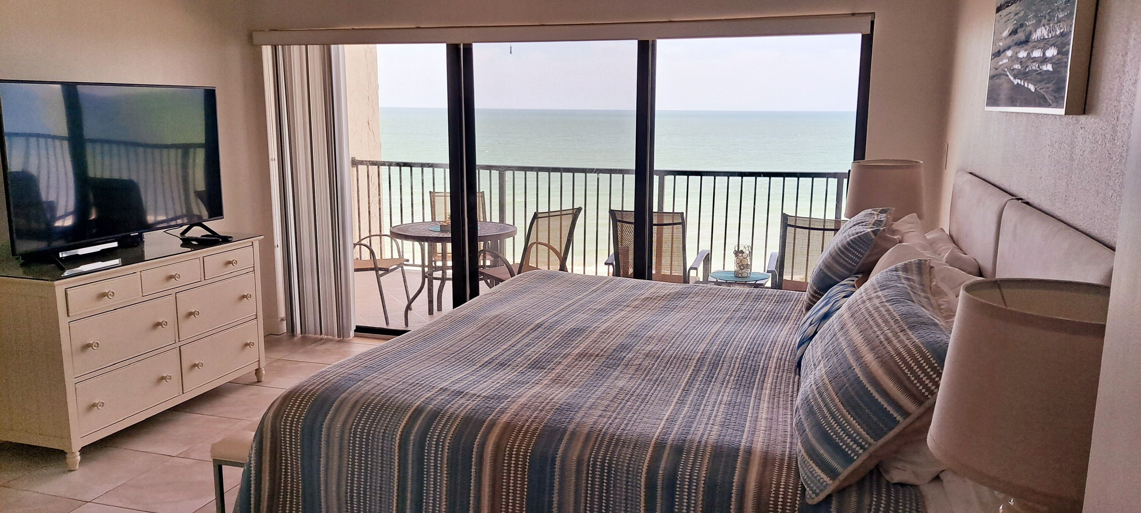 Una habitación de hotel con cama, televisión, cómoda, balcón con vista al mar y una pequeña mesa con sillas.