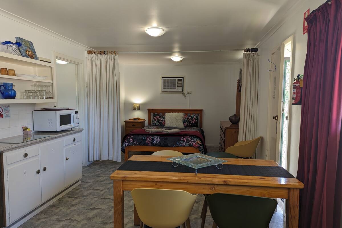 Een compacte slaapkamer met een bed, nachtkastje en een klein bureau met stoelen. Er staat een magnetron op het aanrecht en er is een raam met gordijnen.