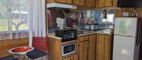 A compact kitchen with wooden cabinets, a built-in microwave, a refrigerator, and a small dining area with a glass table and red chairs.