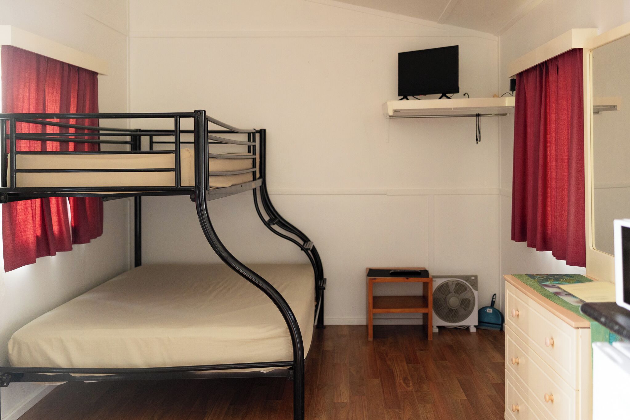Une chambre avec un lit superposé, un téléviseur fixé au-dessus, un petit ventilateur et une commode avec un miroir.
