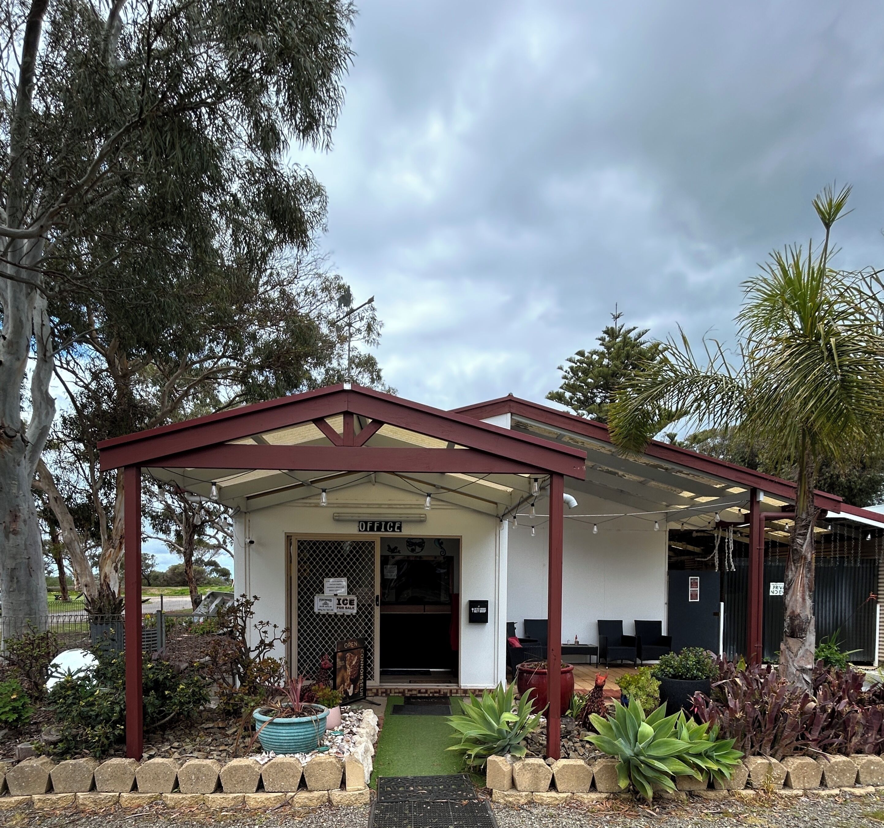 A building with a covered entrance labeled 'OFFICE,' surrounded by various plants and trees.