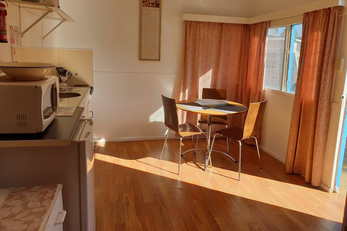 Een kleine keuken met een magnetron, een kom en een spoelbak. Er is een eettafel met stoelen en een raam met gordijnen.