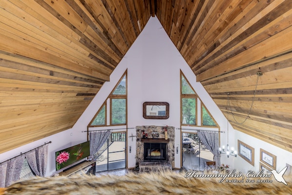A cabin interior with a high ceiling, wooden beams, a fireplace, and large windows.