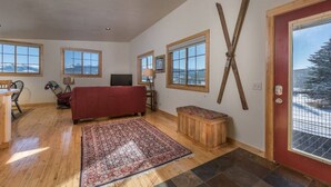 A spacious room with wooden flooring, a red sofa, a patterned rug, and large windows offering a view of snow-covered mountains. - Unique Driggs Quiet Apartment (Driggs)