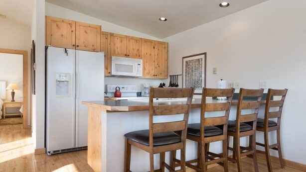 A modern kitchen with wooden cabinets, a white refrigerator, a microwave, and a kettle. There are four wooden chairs around a small island with a sink. - Unique Driggs Quiet Apartment (Driggs)