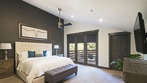 Un dormitorio con una cama grande, un ventilador de techo, un ventanal corredero a un balcón y un televisor montado en la pared.