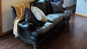 A cozy living room with a black leather sofa, a herringbone throw blanket, and patterned pillows. A round mirror with a seascape hangs on the wall, and a window with dark curtains allows natural light.