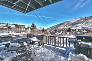 A balcony with a metal grill and a table, overlooking a snowy landscape with houses and a hill.