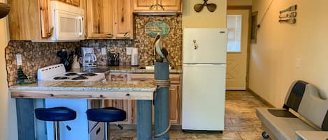 A compact kitchen with wooden cabinets, a central island with stools, a refrigerator, and a stone backsplash.