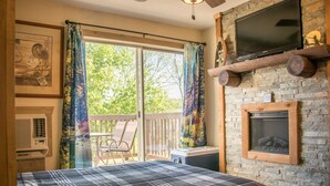 A bedroom with a bed, a TV mounted on a stone wall, a fireplace, and a balcony with a chair and table.