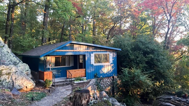A small blue cabin with a stone wall foundation, a wooden bench, and a window overlooking a forested area.