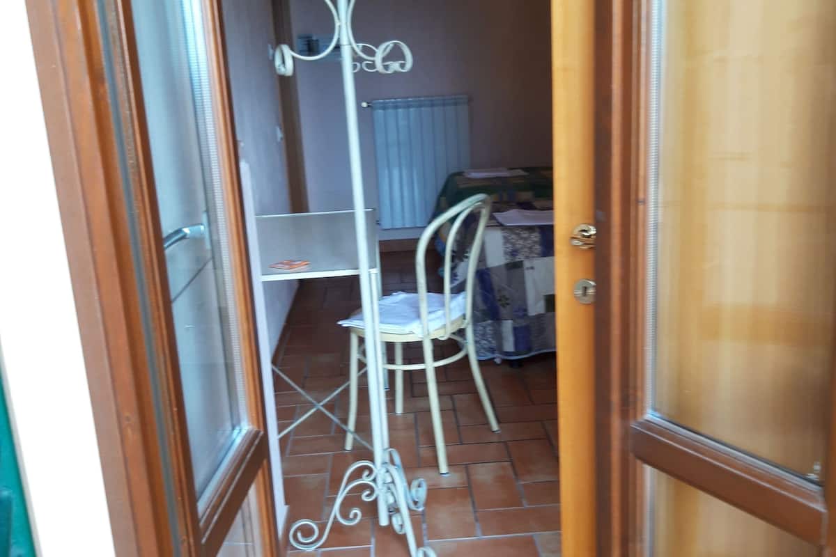 A narrow hallway with a tiled floor, a white metal chair, a radiator, and a bed with a green and white bedspread.