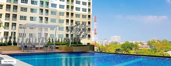 A swimming pool with a sunlit outdoor seating area, a modern building with balconies, and a clear view of the surrounding cityscape.