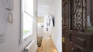 A hallway with a wooden floor, a radiator, a white door with a brass handle, and a window with blinds.