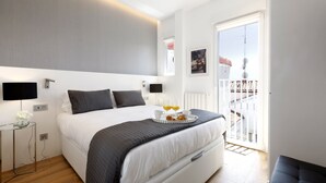 A modern hotel room with a large bed, bedside tables, a glass coffee table, and a balcony with a railing.