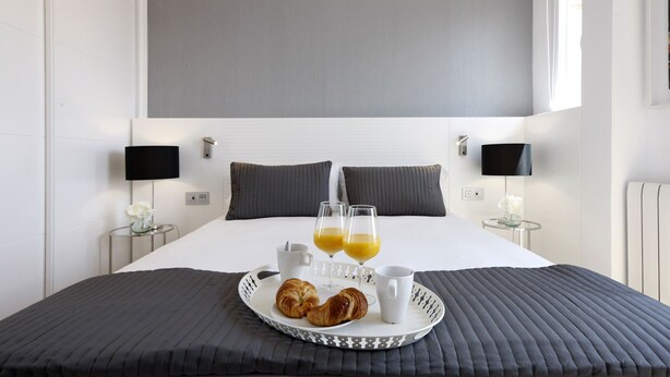 A neatly made bed with a tray of croissants, two glasses of orange juice, and a cup, set against a modern room with a large headboard and bedside lamps.