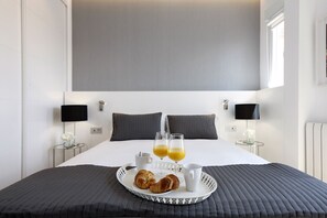 Una cama bien hecha con una bandeja de cruasanes, dos vasos de zumo de naranja y una taza, frente a un cuarto moderno con un cabecero grande y lámparas de noche.