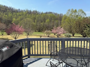 A deck with a grill, a metal table, and chairs, overlooking a grassy area with trees and a forest in the background. - Come for the Mountains. Stay for Everything Else. (Lovingston)