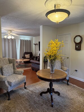 A living room with a patterned armchair, a round wooden table with a vase of yellow flowers, a ceiling fan, and a wall-mounted clock. - Come for the Mountains. Stay for Everything Else. (Lovingston)