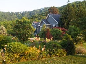 A two-story house with a gabled roof, surrounded by a lush garden with various flowering plants and trees. - Come for the Mountains. Stay for Everything Else. (Lovingston)