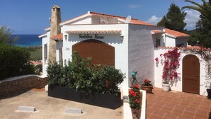 A white building with a red-tiled roof, a sign reading "FantaSea Home," and a view of the sea in the background. - Luxury Villa, Breathtaking 180° Sea & Sunset views, Private Pool. Fully AC (San Jaime Mediterráneo, Son Bou, Menorca)