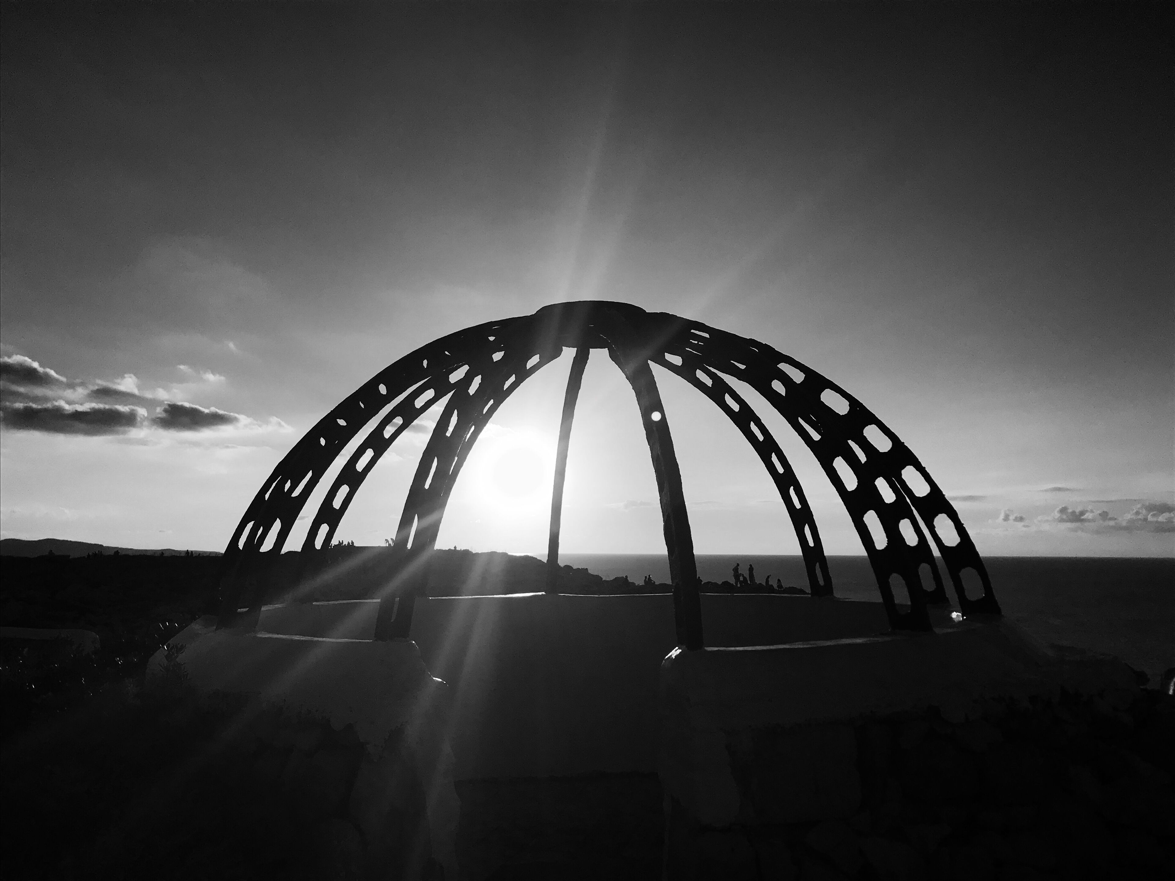 A black and white photo of a large, arched metal structure with multiple openings, set against a backdrop of a clear sky and distant landscape.