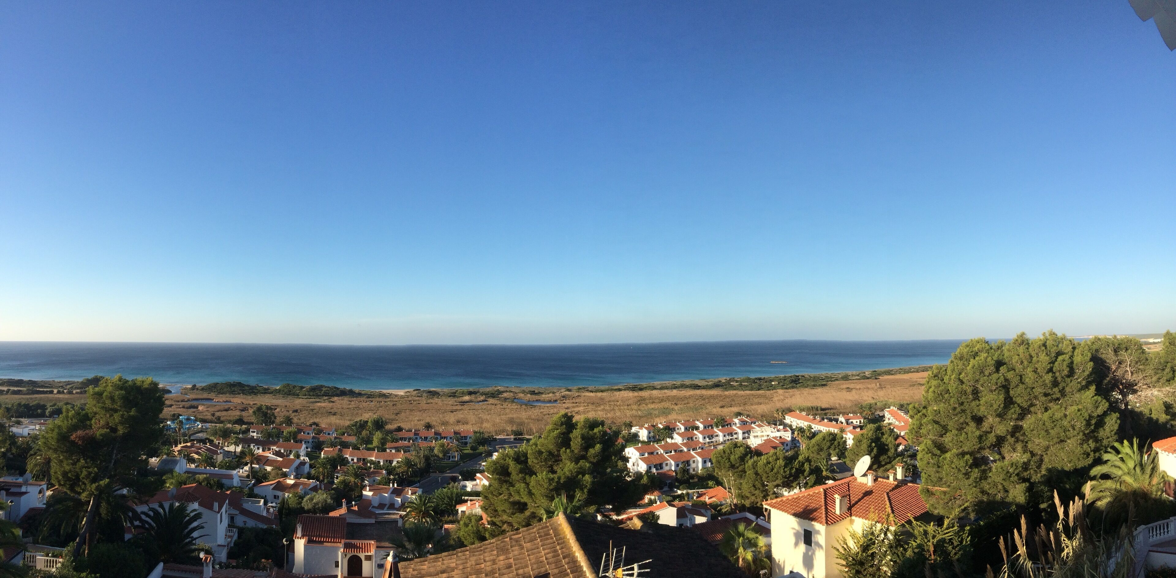 A coastal town with white buildings and red roofs, a clear blue sky, and the sea in the background.