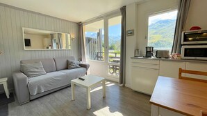 A modern room with a sofa, a small table, a dining table, a kitchen area with a microwave, and a large window with a mountain view.