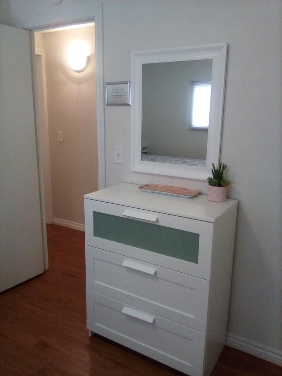 A white dresser with a mirror and a tray, a small plant, and a framed certificate on the wall.
