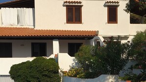 A white building with a tiled roof and brown shutters, surrounded by a landscaped garden with steps and a small table with an umbrella.