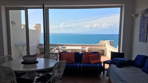 A room with a dining table, chairs, a sofa, and a view of the sea.