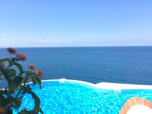 Une piscine à l’eau d’un bleu cristallin, entourée d’une terrasse blanche, et offrant une vue imprenable sur l’océan sous un ciel dégagé.