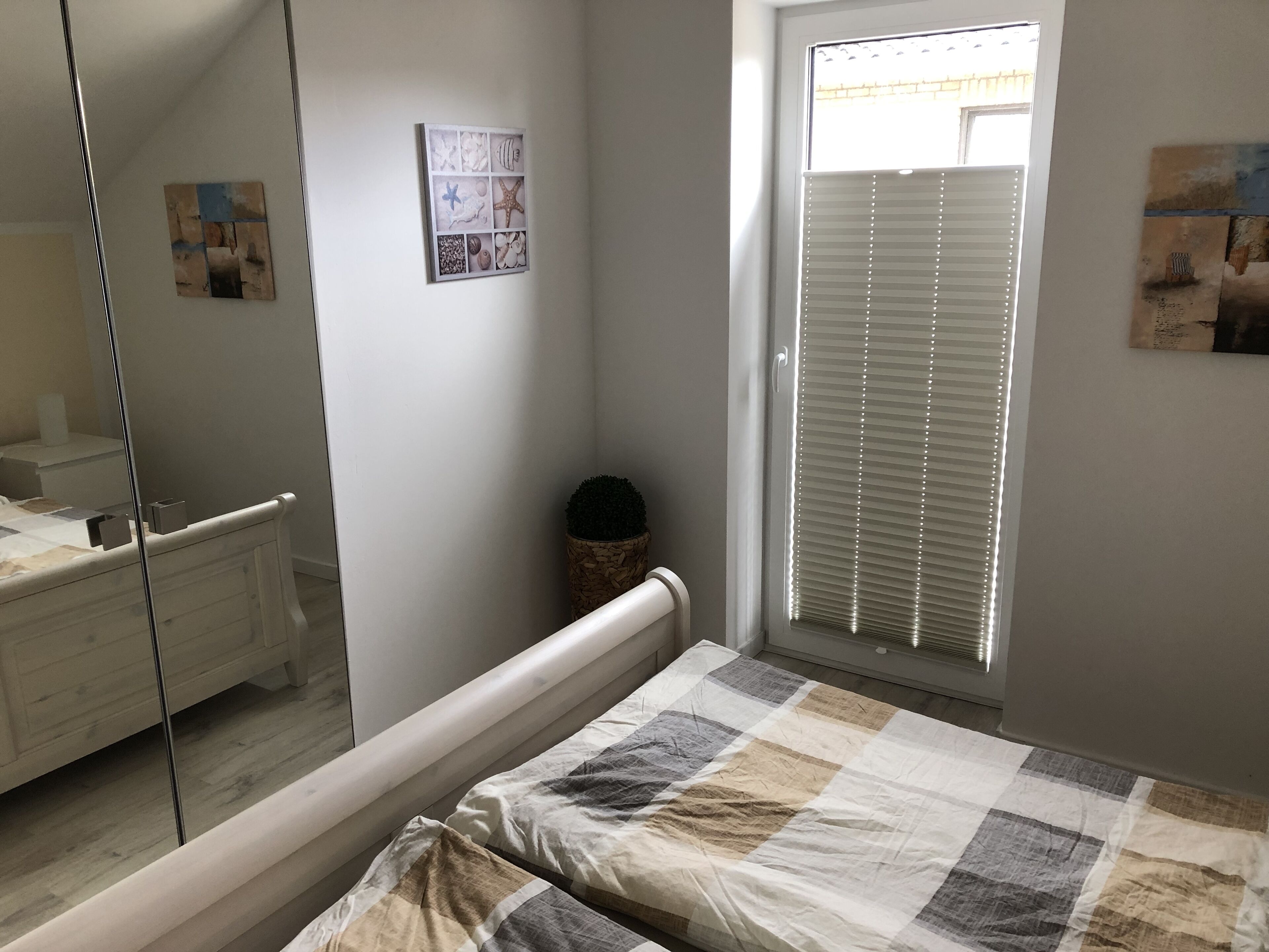 Ett sovrum med en säng, ett sängbord, en spegel, ett fönster med persienner och en vägg med en inramad tavla.