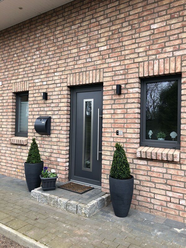 A modern brick building with a dark grey door, potted plants, and a window with decorative elements.