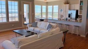 A spacious living room with a white sofa, a coffee table, and a view of the beach through large windows. - Just Steps to the Ocean, Beachfront, House On the Beach, Minutes from Galveston (Freeport)
