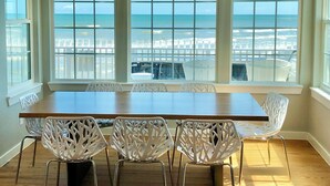 A dining area with a large table and white chairs, large windows with a view of the ocean, and a ceiling light fixture. - Just Steps to the Ocean, Beachfront, House On the Beach, Minutes from Galveston (Freeport)