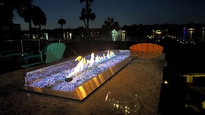 Ein Feuerstelle mit Glaseinfassung, eine Sitzgruppe mit Stühlen und im Hintergrund Blick auf Wasser und Stadtlichter.