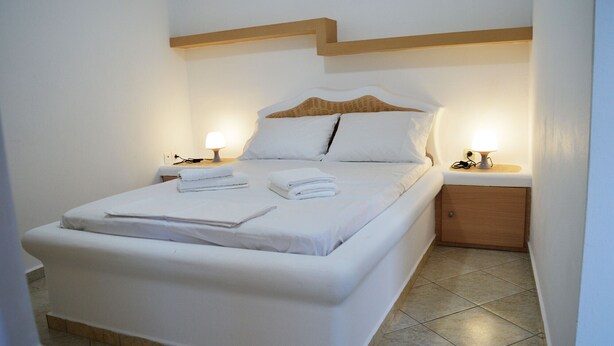 A neatly made bed with white linens and towels, two bedside lamps, and a wooden nightstand with a lamp. - Apartment Danai is situated near the Centre of Ermoupolis (Ano Siros)