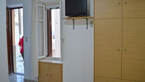 A room with a television on a wooden shelf, a window with blinds, and wooden wardrobes. - Apartment Danai is situated near the Centre of Ermoupolis (Ano Siros)