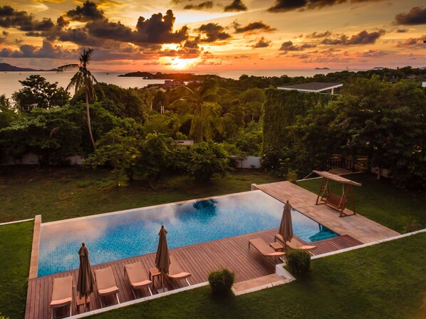 Une piscine à débordement avec une terrasse en bois et des chaises longues, entourée d’une végétation luxuriante et offrant une vue imprenable sur le coucher de soleil.