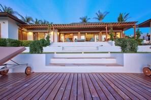 Una casa moderna con terraza de madera, escalones que conducen a un espacio exterior cubierto y palmeras al fondo.