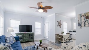 Ein Wohnzimmer mit blauem Sofa, Fernseher, Deckenventilator und einem Essbereich mit Tisch und Stühlen.