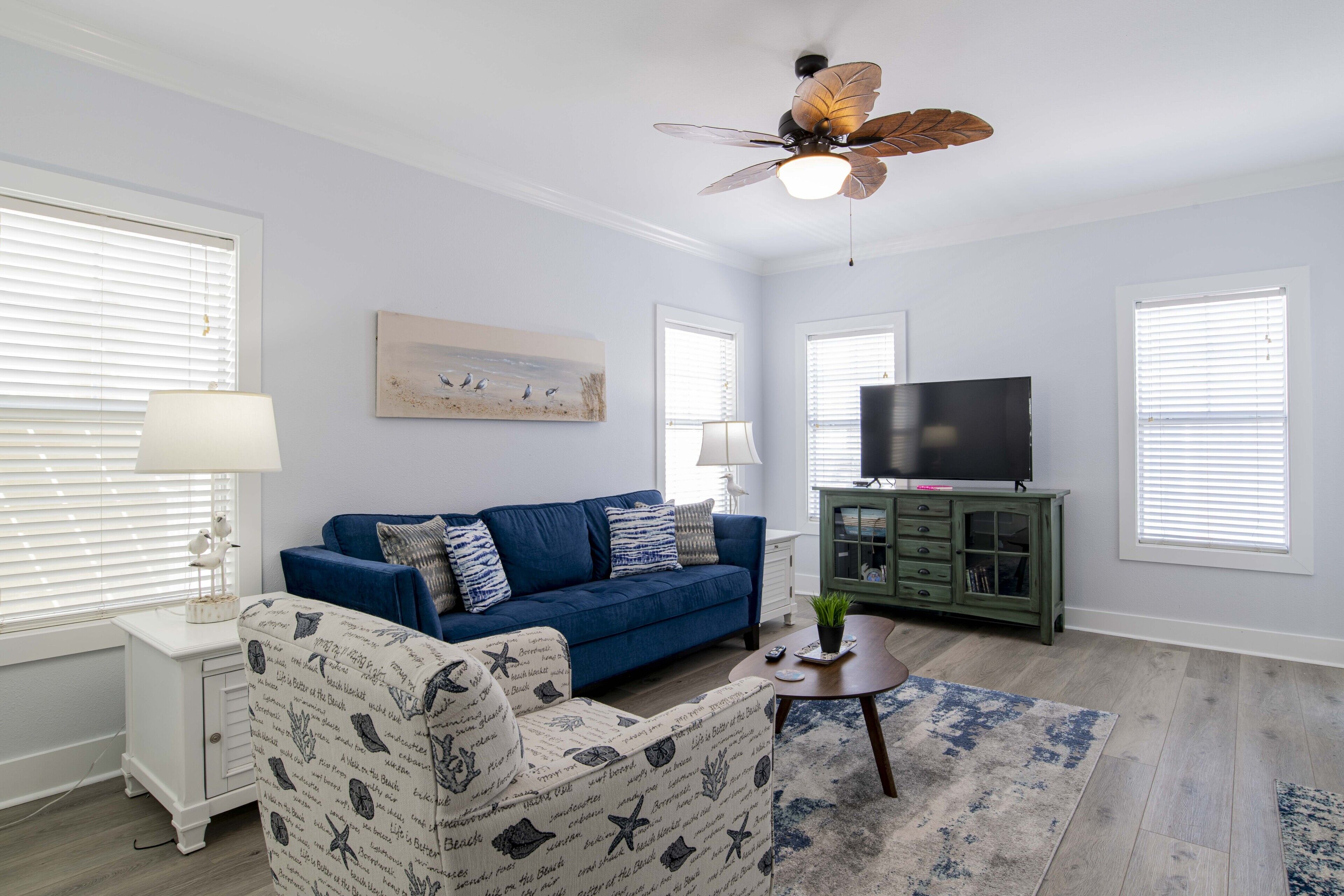 Un salon avec un canapé bleu, un fauteuil blanc, un ventilateur de plafond, un téléviseur à écran plat et un tableau accroché au mur.