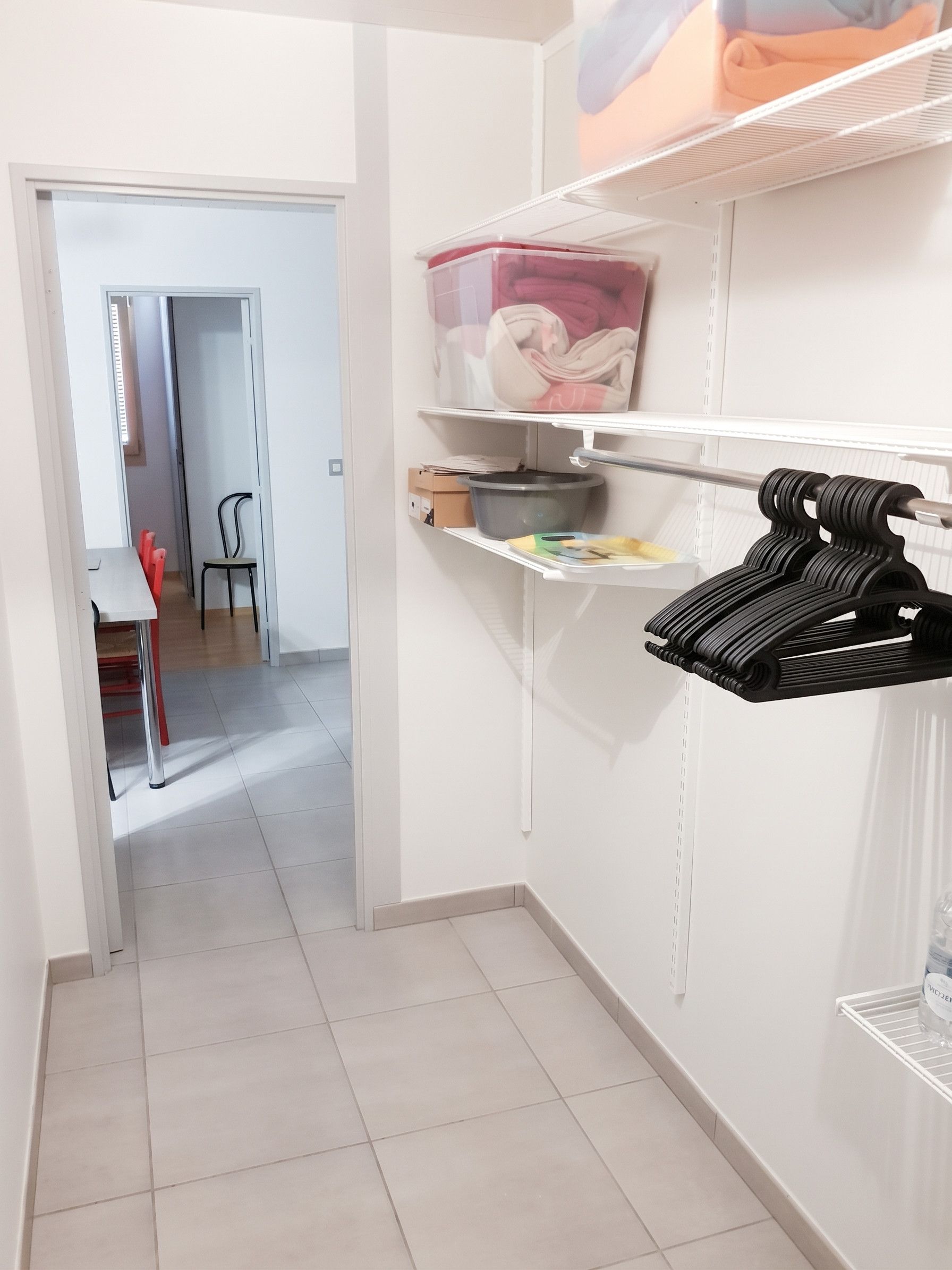 Un couloir étroit, avec une porte blanche au fond, une étagère blanche sur laquelle se trouve un récipient en plastique, et un présentoir noir contenant divers objets.