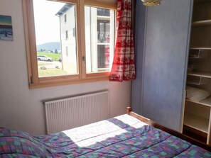 A bedroom with a bed, a radiator, a window with a view of a house and cars, and a wardrobe. - Homerez - Apartment – Métabief (Métabief)