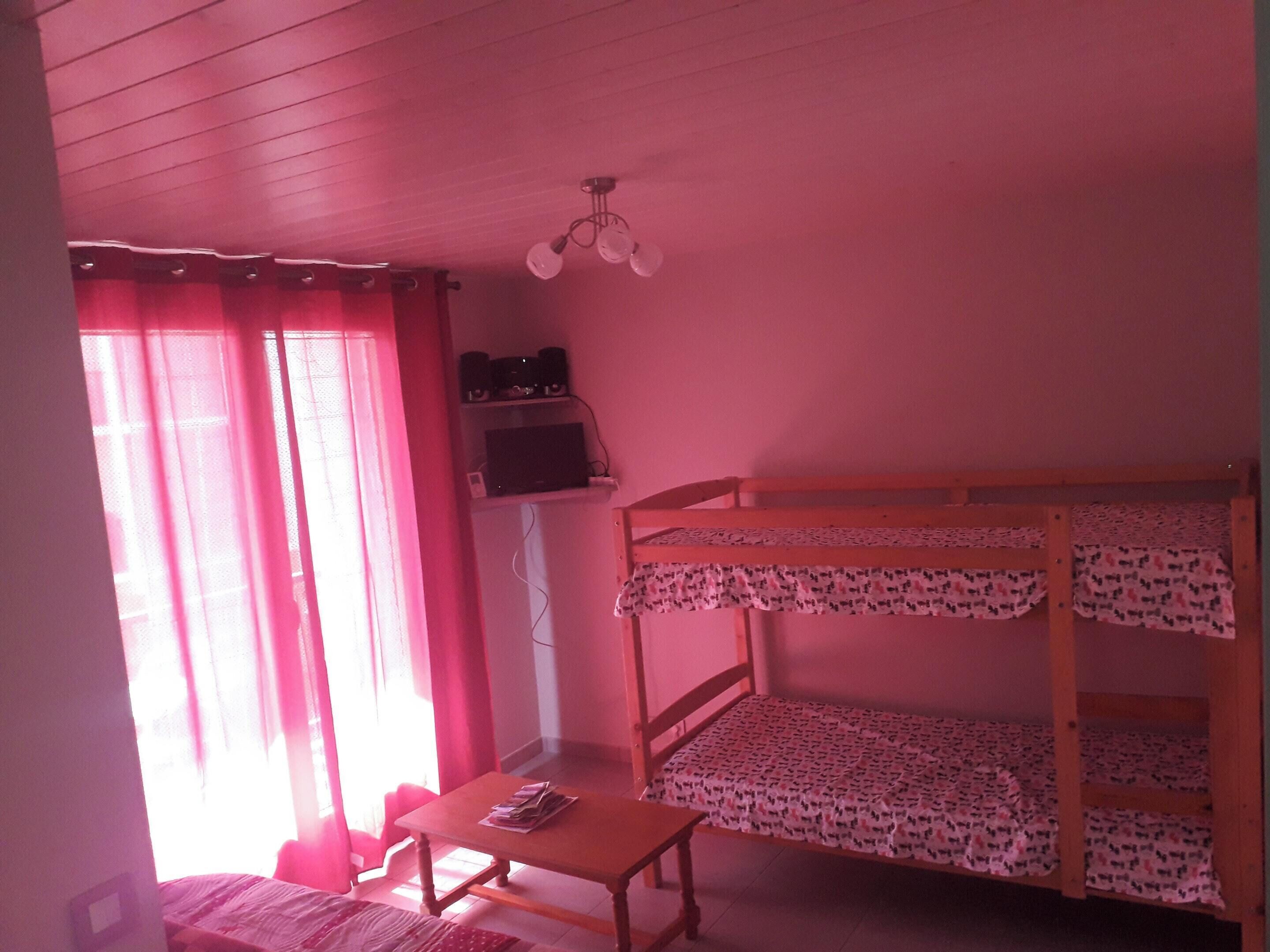 Una habitación con literas, una mesita pequeña, una estantería con altavoces y una ventana con cortinas.