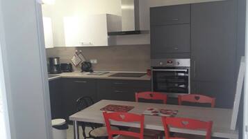 Ett kompakt kök med moderna vitvaror, ett matbord med stolar och en synlig papperskorg.