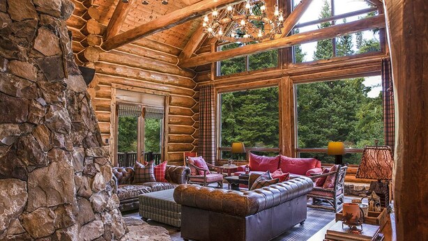A log cabin interior with a stone wall, wooden beams, large windows, and a cozy seating area with a fireplace. - Limber Pine Lodge by Cottonwood Lodging (Brighton)
